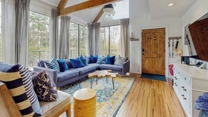 A spacious living room with a large sofa, a wooden coffee table, and a view of the outdoors through large windows. - Dog-OK, updated home with WiFi, Sauna,  game room - close to lakes & hiking (Lake Arrowhead)
