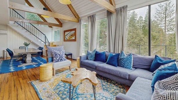 A spacious living room with a wooden ceiling, a gray sofa with blue cushions, a dining area with a wooden table, and a staircase leading to an upper level. - Dog-OK, updated home with WiFi, Sauna,  game room - close to lakes & hiking (Lake Arrowhead)