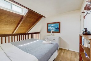 A bedroom with a sloped ceiling, a bed with a blue and white bedspread, a nightstand with a lamp, a framed picture on the wall, and a bookshelf with books.