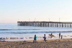A beach with a pier extending into the water, people walking along the shore, and a child with a surfboard. - Charming, two-story home with deck, grill, and ocean views (Cayucos)