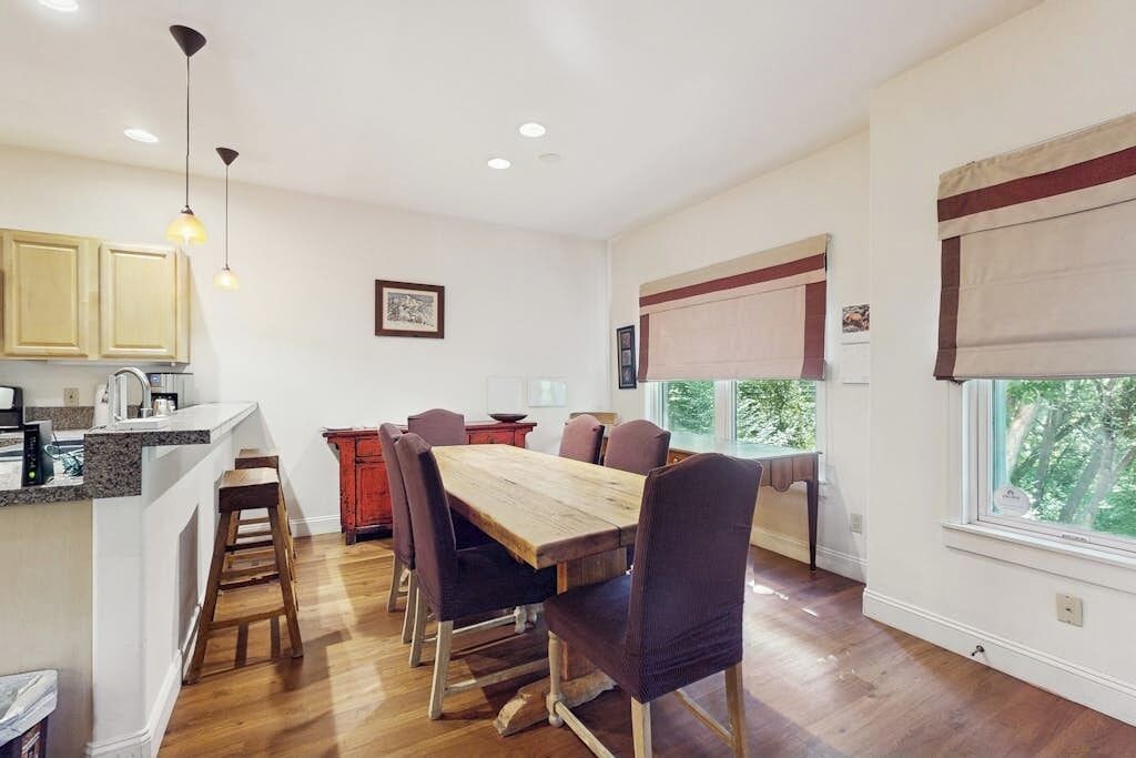 A dining area with a wooden table and chairs, a kitchen with cabinets, and a window with a view of trees.