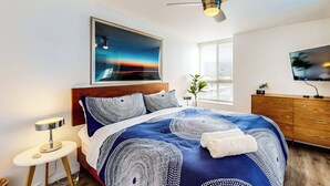 Een slaapkamer met een bed, een plafondventilator, een nachtkastje, een televisie en een raam met jaloezieën.