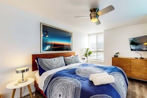A bedroom with a bed, a ceiling fan, a nightstand, a TV, and a window with blinds. - Contemporary Condo | Mountain Views & Balcony | 1 Block to Beach (Carpinteria)