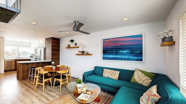 Een moderne woonruimte met een eethoek, een blauwgroene bank, een glazen salontafel en een groot schilderij van een zonsondergang.