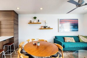 A modern living room with a round wooden table, a teal sofa, and a ceiling fan. - Contemporary Condo | Mountain Views & Balcony | 1 Block to Beach (Carpinteria)