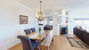 Uma sala de estar ampla com uma mesa de jantar de madeira, cadeiras estofadas cinzas, um sofá cinza, uma lareira e um candelabro.