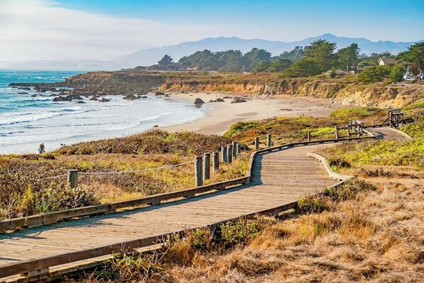 A coastal walkway leading to a beach with a view of the ocean and distant mountains.