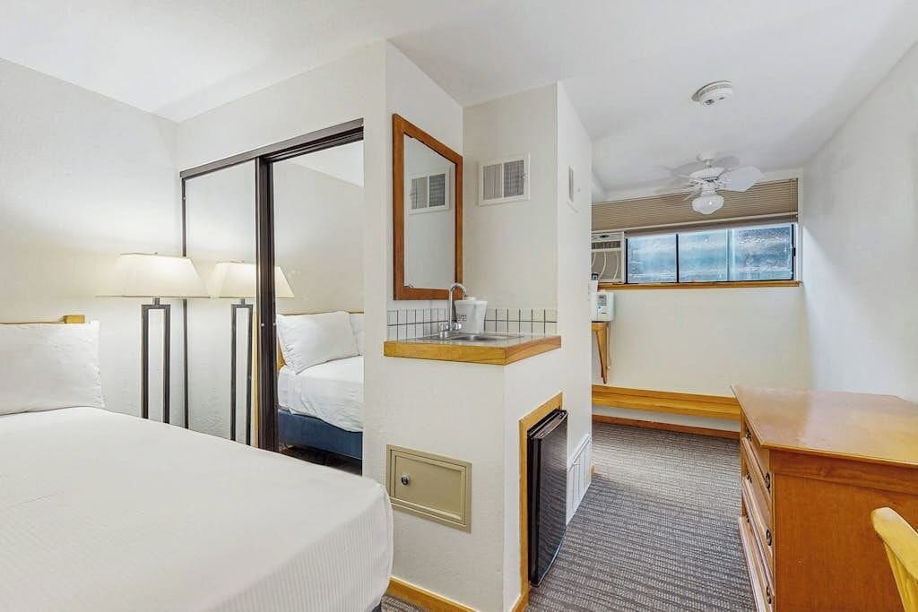 Una camera d'albergo con un letto, una scrivania, uno specchio, una finestra e una ventola a soffitto.