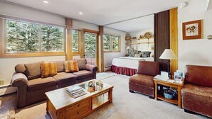 Ein geräumiges Wohnzimmer mit einer Couch, einem Sessel und einem Couchtisch, große Fenster mit Blick auf Bäume und ein Bett mit roter Bettdecke.