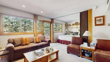 Un salon spacieux avec un canapé, un fauteuil et une table basse, de grandes fenêtres donnant sur la verdure, et un lit recouvert d’une courtepointe rouge.