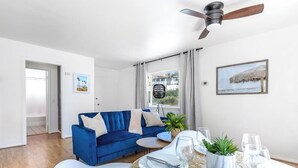 Ein Wohnzimmer mit blauem Sofa, einem Esstisch gedeckt mit Tellern und Gläsern, einem Deckenventilator und einem Fenster mit Vorhängen.