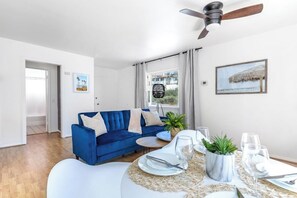 Ein Wohnzimmer mit blauem Sofa, einem Esstisch gedeckt mit Tellern und Gläsern, einem Deckenventilator und einem Fenster mit Vorhängen.