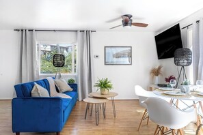 A modern living room with a blue sofa, a dining table with chairs, a ceiling fan, a TV, and a window with curtains.