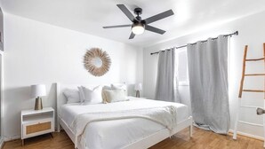 Ein Schlafzimmer mit Bett, Deckenventilator, Nachttisch, Lampe, Wanddekoration und einem Fenster mit Vorhängen.