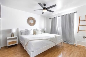 Ein Schlafzimmer mit Bett, Deckenventilator, Nachttisch, Lampe, Wanddekoration und einem Fenster mit Vorhängen.