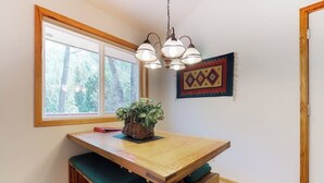 Une pièce comprenant une table en bois, un banc rembourré de coussins verts, un luminaire suspendu, une fenêtre avec vue et un objet décoratif fixé au mur.