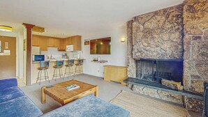 Un salon moderne avec une cheminée en pierre, une cuisine avec des meubles en bois et un coin bar avec des tabourets.
