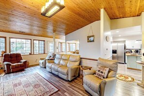Ein geräumiges Wohnzimmer mit Holzboden, einer Sofagarnitur, einem Teppich und einem durch eine offene Durchgangstür einsehbaren Küchenbereich.
