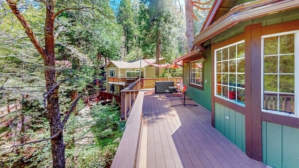 Una terraza de madera con vista a una zona boscosa, un porche cubierto con mobiliario exterior y un edificio con grandes ventanales.