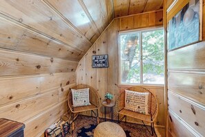 Ein Blockhaus-Interieur mit Holzwänden, zwei Korbstühle und Schildern mit See-Regeln, ein kleiner Tisch mit einer Pflanze und ein Fenster mit Blick auf Bäume.