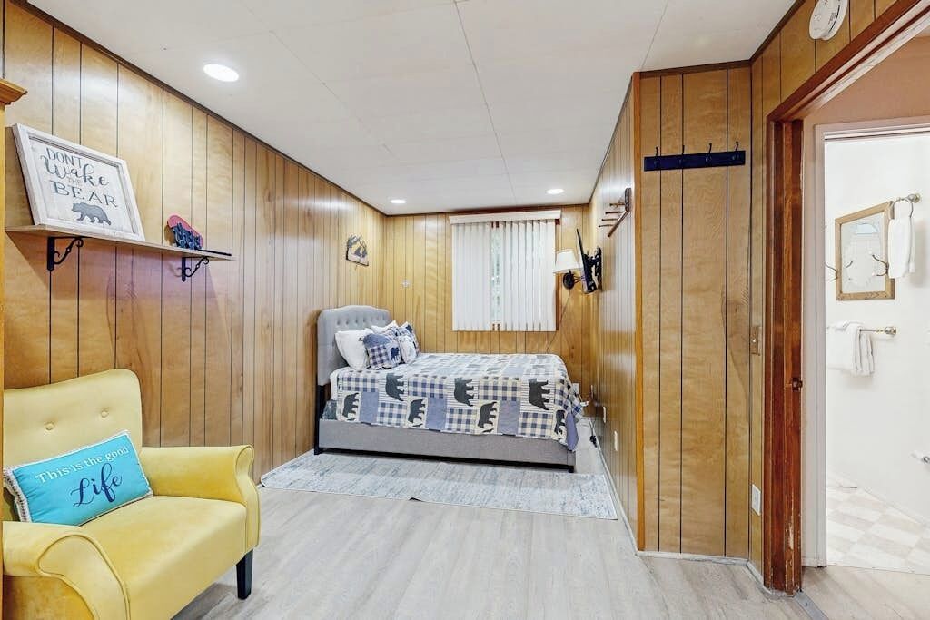 A bedroom with a bed, a yellow armchair, wooden paneling, a clock, and a door.