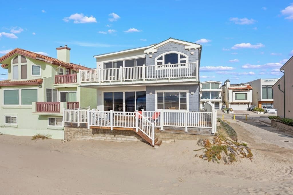 Une maison à deux étages avec un balcon, un porche à l’avant et une allée bordée de sable.