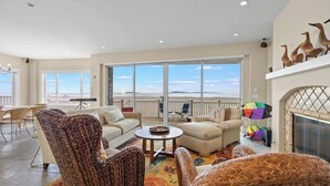 A spacious living room with a large window overlooking a scenic view, a fireplace, and wicker furniture. - Beachfront abode with spacious deck, pool table, gas grill, smart TVs, & W/D (Oxnard)