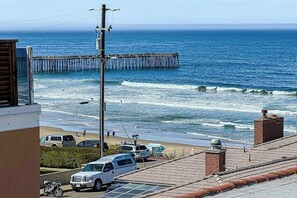 A coastal view with a pier extending into the ocean, a beach with parked cars, and a building with a rooftop. - Beautiful Oceanview home with outdoor grill and spacious rooftop (Pismo Beach)