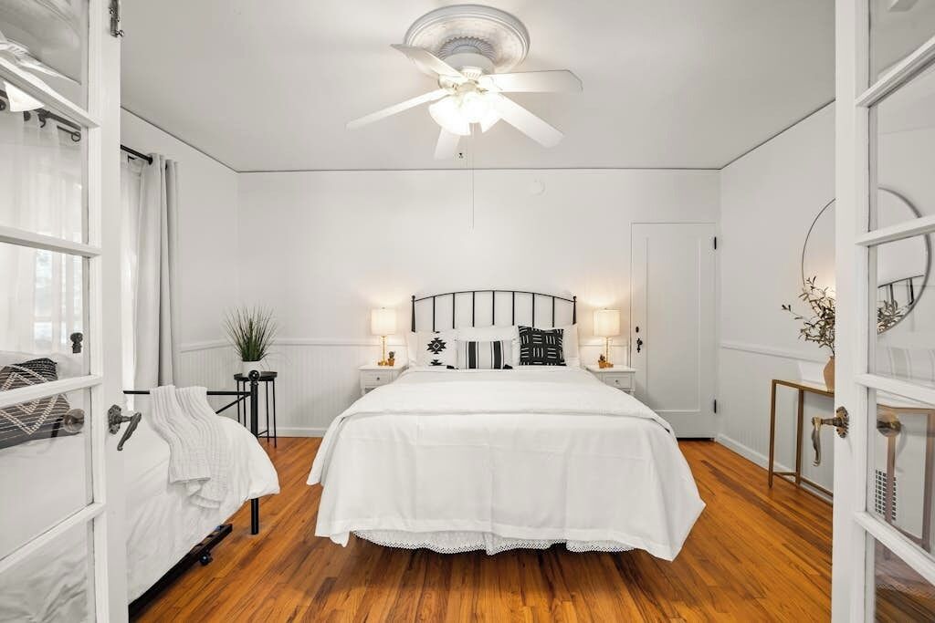 A bedroom with a bed, a ceiling fan, bedside lamps, a nightstand, a potted plant, and a door.