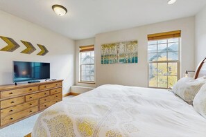 Ein Schlafzimmer mit Bett, Kommode, Fernseher, Fenstern und Kunstwerken.