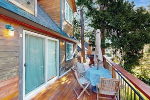 A wooden deck with a table and chairs, a covered patio area, and a view of trees. - Mountain-view cabin with fireplace, outdoor dining, firepit & wraparound deck (Lake Arrowhead)