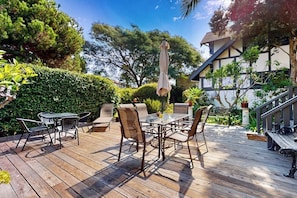 A wooden deck with a glass-top table and chairs, surrounded by greenery and a building with a balcony.