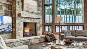 Une cheminée en pierre, une grande fenêtre donnant sur les arbres et un coin salon confortable avec une table basse en verre.