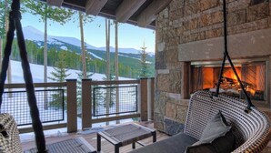 Un espace extérieur accueillant, avec un coin salon, une cheminée en pierre, un fauteuil suspendu et une vue sur des montagnes enneigées.
