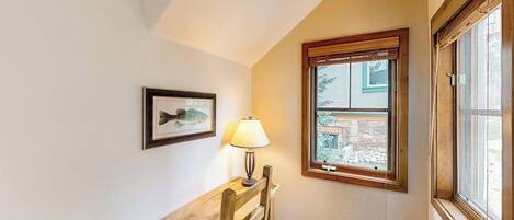 A corner room with a wooden bench, a framed picture, a lamp, and a window with a view of a stone building.