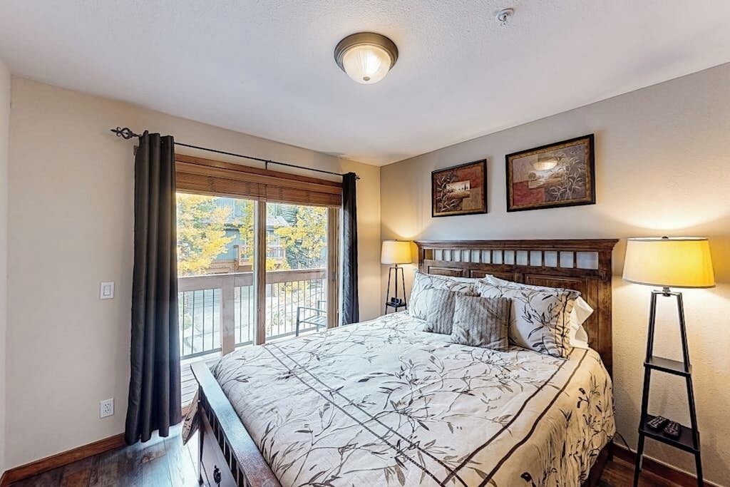 Un dormitorio con una cama grande, dos lámparas de noche, una ventana con cortinas y dos cuadros en la pared.
