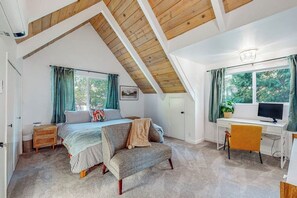 Ein Schlafzimmer mit schrägem Dach, einem Bett, einem Schreibtisch, einem Stuhl und einem Fenster mit Vorhängen.
