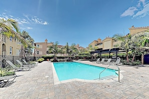 Ein Swimmingpool mit Liegestühlen und Sonnenschirmen, umgeben von Gebäuden und Palmen.