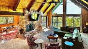 A spacious living area with a fireplace, a green sofa, and a dining table with chairs. - 3BR/2.5BA home with views, yard, BBQ, game room, fireplace & EV charger (Lake Arrowhead)