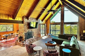 A spacious living area with a fireplace, a green sofa, and a dining table with chairs. - 3BR/2.5BA home with views, yard, BBQ, game room, fireplace & EV charger (Lake Arrowhead)