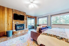 A room with a fireplace, a television, a sofa, a bed, and large windows. - 3BR/2.5BA home with views, yard, BBQ, game room, fireplace & EV charger (Lake Arrowhead)