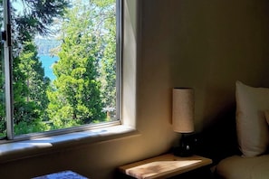 Ein Zimmer mit Fensterblick auf Wald und Gewässer, einem Nachttisch mit Lampe und einem Bett mit Kissen.