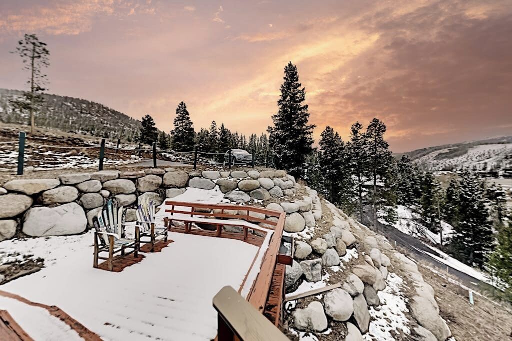 A snow-covered outdoor seating area with stone walls and wooden benches, surrounded by trees and a view of a hilly landscape.