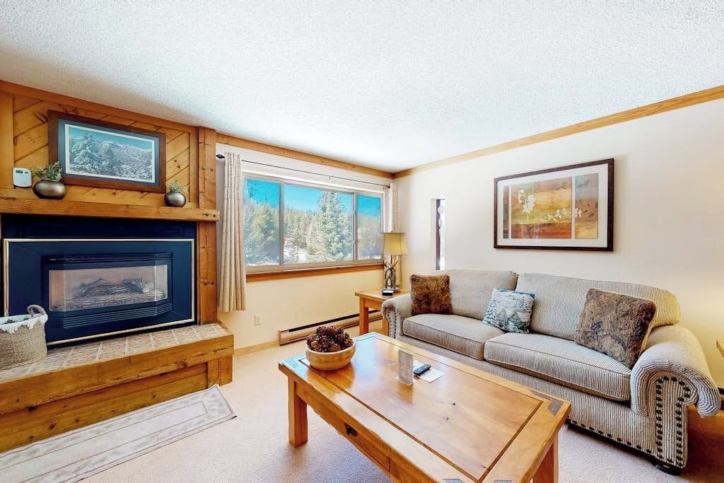 A living room with a fireplace, a sofa, a coffee table, and a window with a view of trees.