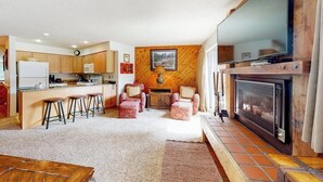 A kitchen with wooden cabinets, a dining area with stools, a fireplace, and a large TV mounted on a wooden wall. - Dog-friendly ski condo with hot tubs, grills, & gym (Breckenridge)