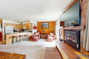 Eine Küche mit Holzschränken, ein Essbereich mit Hockern, ein Kamin und ein großer Fernseher an einer Holzwand.