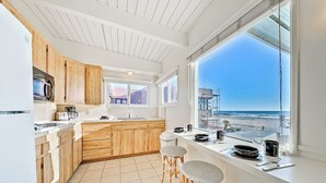 Une cuisine moderne avec des meubles en bois, une grande fenêtre donnant sur la plage et deux tabourets de bar.
