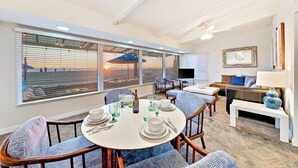 Un coin repas avec une table ronde dressée pour six personnes, de grandes fenêtres équipées de stores, un canapé et un téléviseur.