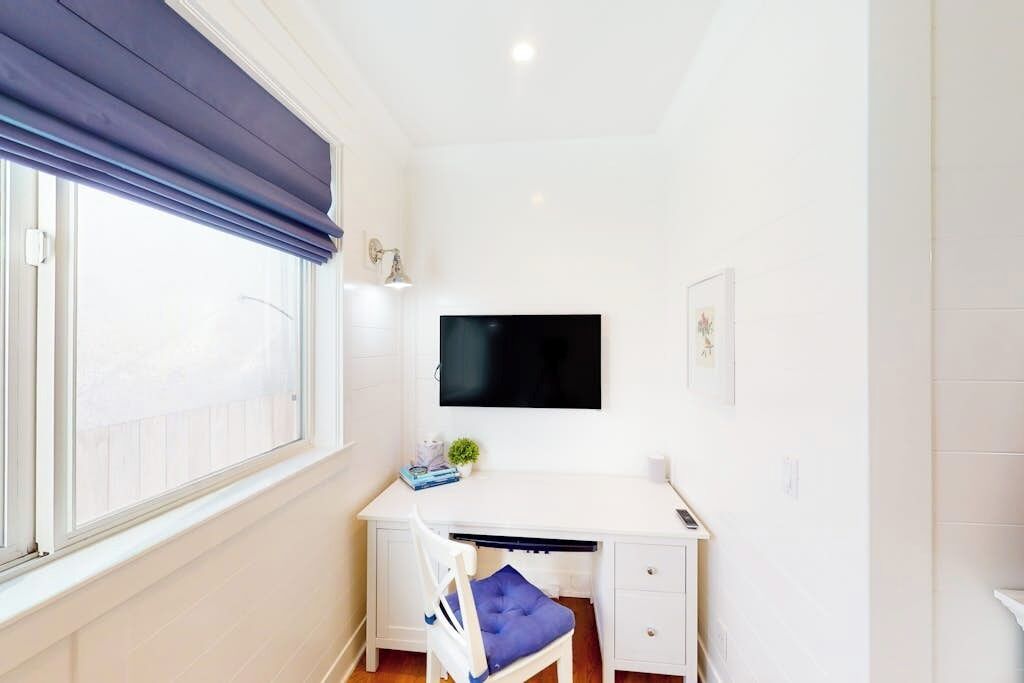 Quarto branco com escrivaninha, cadeira e televisão na parede.