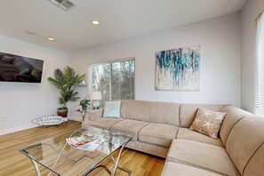 A living room with a beige sofa, a glass coffee table, a TV, a large window, and a wall art piece. - Dual homes with 2 full kitchens, fast WiFi, fireplace, & patio - dog-friendly (Newport Beach)